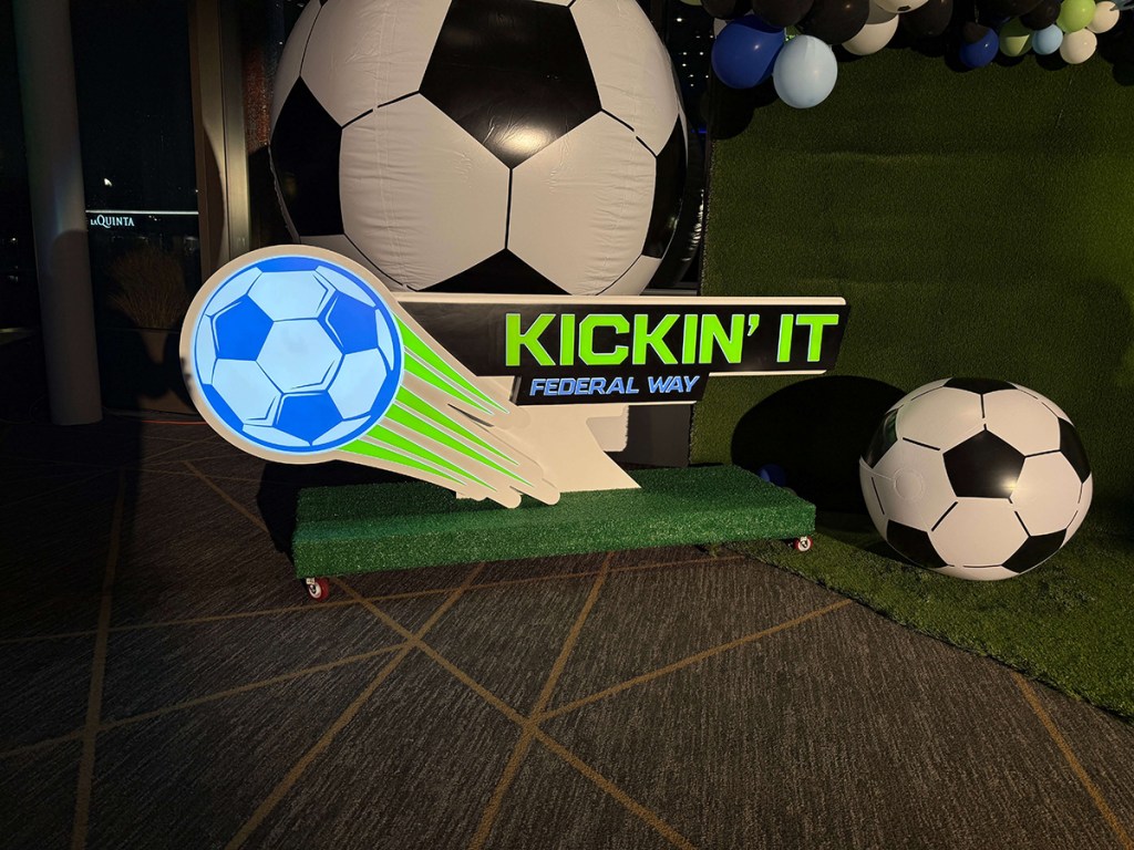 A large “Kickin’ It Federal Way” event display featuring an illuminated sign with a blue and green soccer graphic. Two oversized inflatable soccer balls sit behind and beside the sign, with green turf and balloon decorations creating a sporty, festive backdrop.